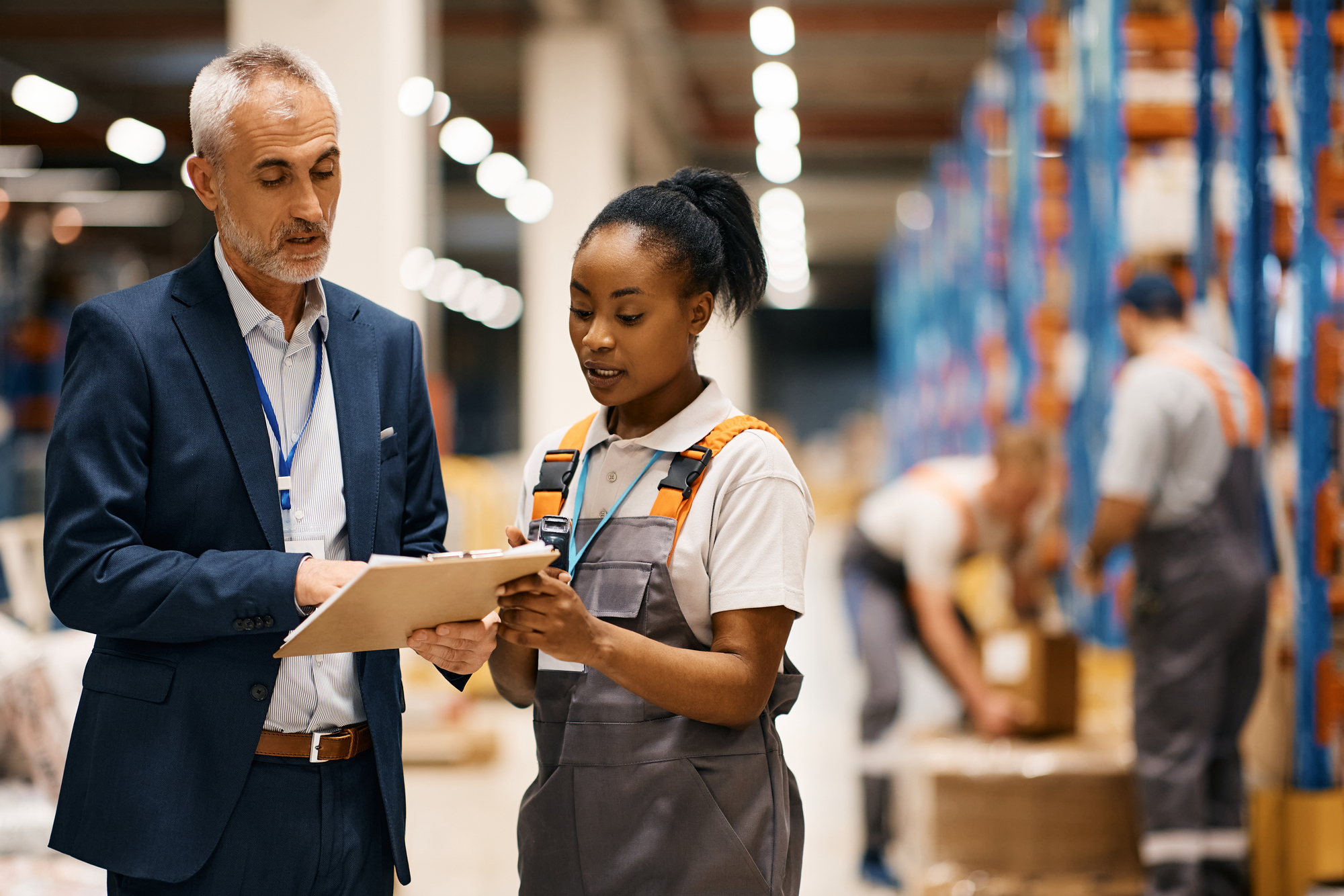 African American warehouse worker and her supervisor cooperating while analyzing reports at storage compartment.