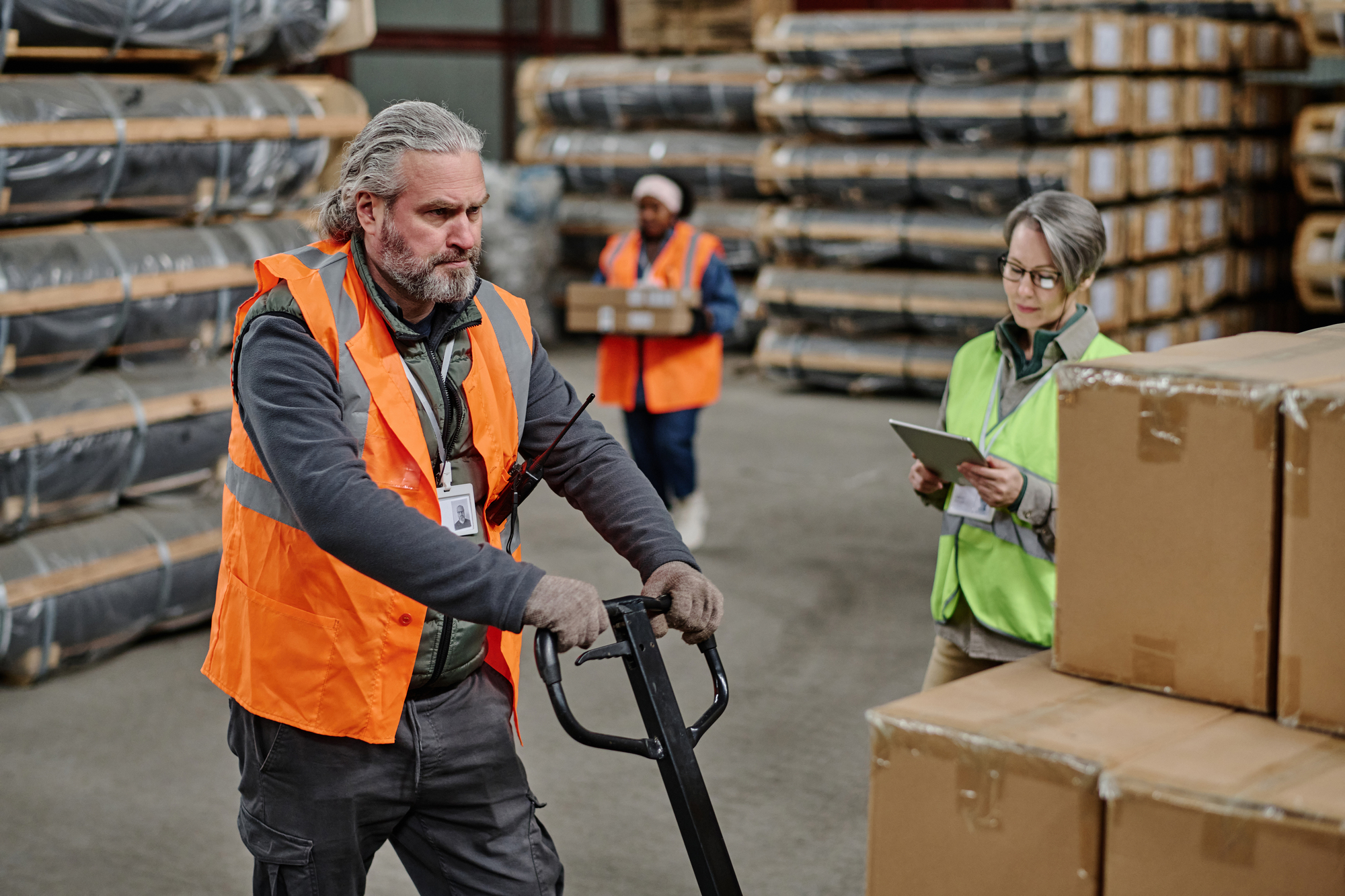 Mature worker in protective workwear loading cargo with loader in storage room with foreman using tablet pc in background